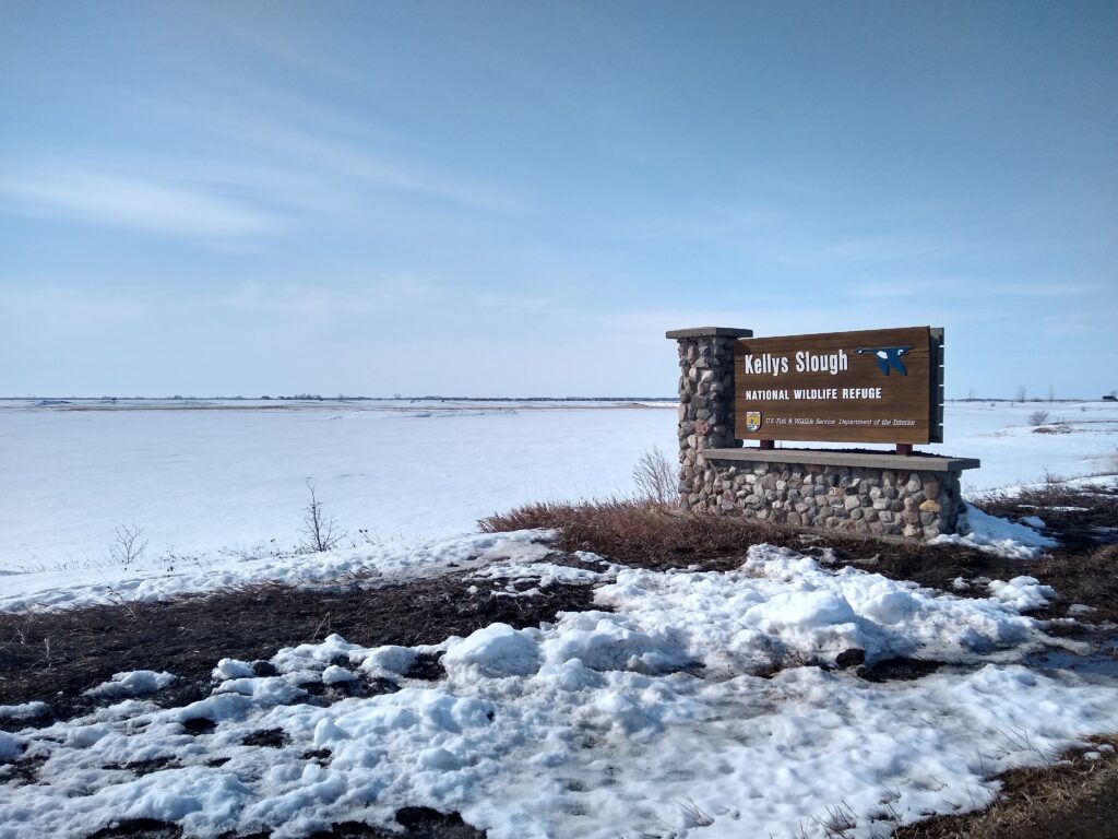 Photo of the sign for "Kellys Slough National Wildlife Refuge" with a snow-covered lake in the background.