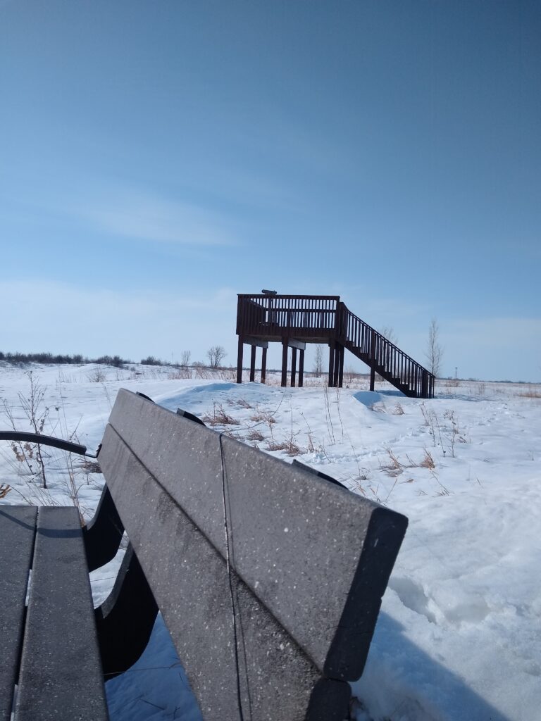 Photo of a bench (foreground) with a wire antenna leading up to an elevated platform (background). There is snow on the ground.