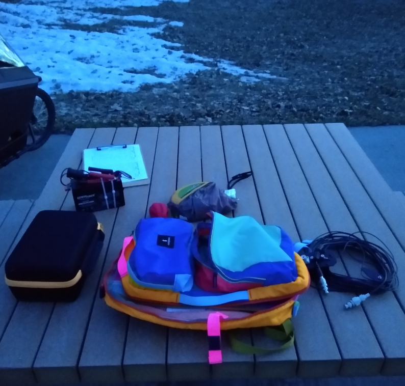 A picnic table at dusk. There is a brightly colored backpack, a black case, a wire antenna, a battery, and a clipboard on it. Some grass and snow are visible in the background. One wheel of a cargo bike is seen in the corner.