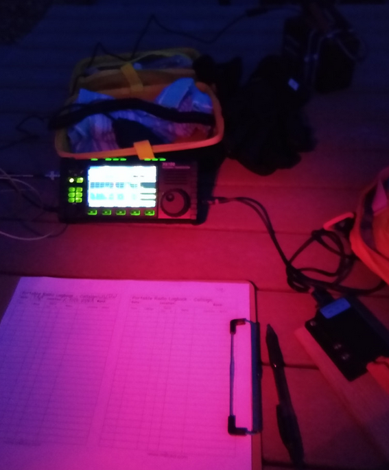 Photo of a picnic table with radio equipment on it. There is a red glow cast by my headlamp. A clipboard and paddle key are in the foreground, with a blurry X6100 display in the back.
