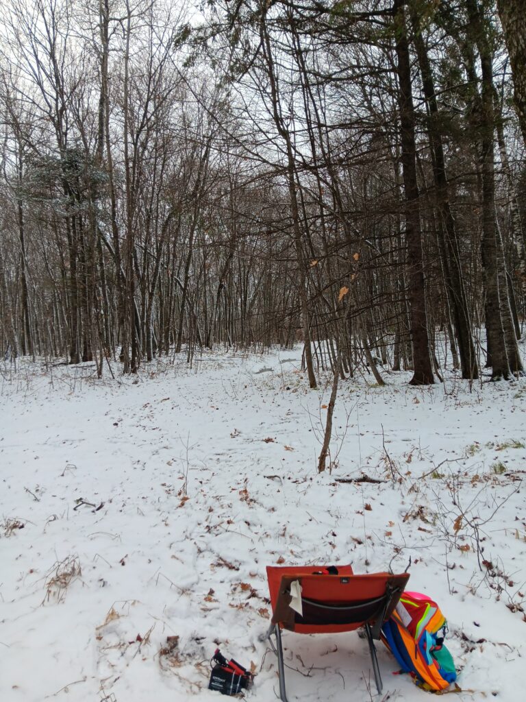 Obligatory gear photo with only the antenna (barely) and battery visible. There is a low orange camp chair in the foreground with a brightly colored backpack leaning on it. A very thin black wire leads from the chair into the trees. Snow on the ground, but still some grass and leaves poking through.