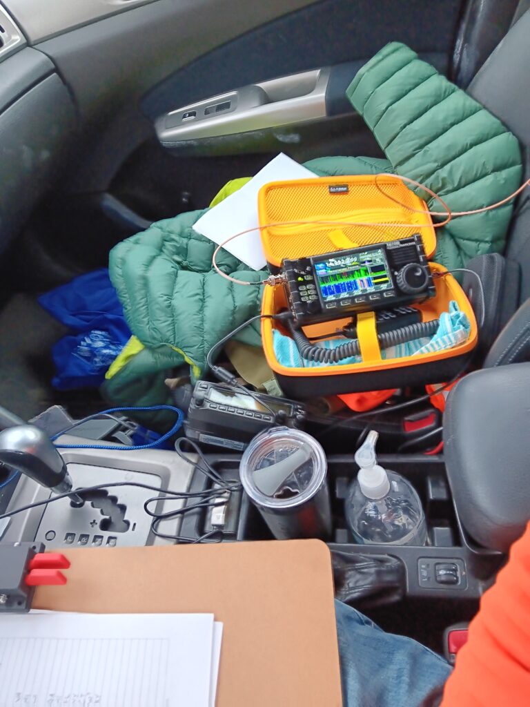 Photo of my messy in-car operating position. Clipboard with key and log, coffee cup, radio propped up on its case, puffy coat and some other stuff underneath that.