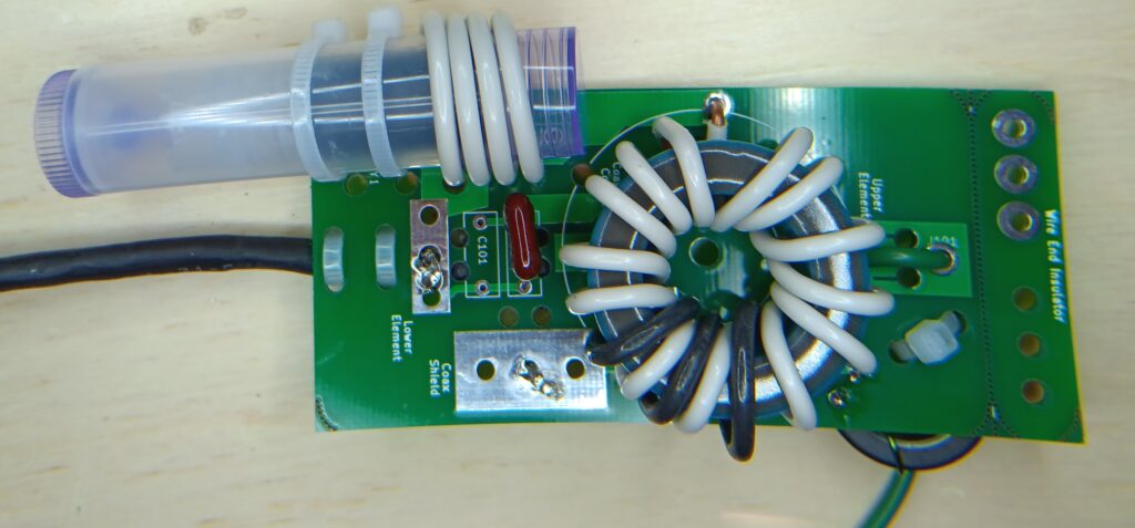 Photo of the assembled Open ARDF 80m Fox Dipole antenna center. There is a toroidal transformer in the middle using two colors of insulated 14 gauge wire (from house wiring) and a variable inductor made from a coil of wire around a lip balm tube, inside of which is a kludged-together ferrite bead. The shrink-wrapped wire is the bottom antenna element and the feedline, and the green wire is the upper antenna element.