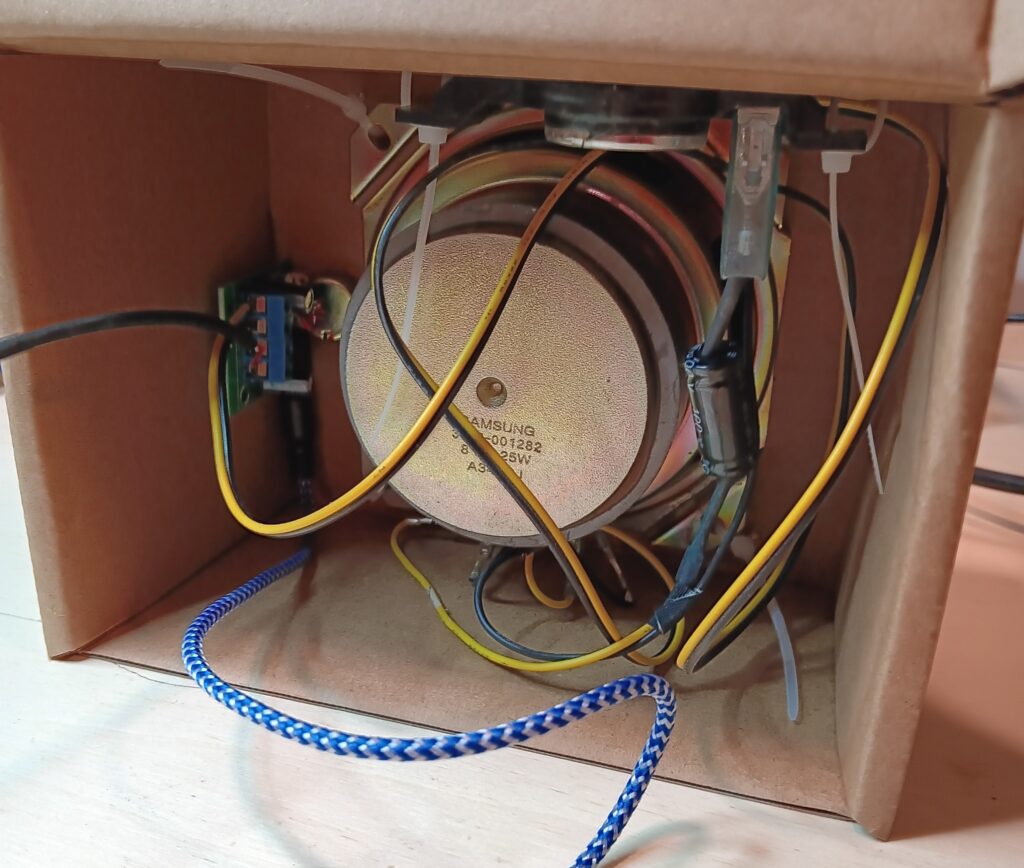 Rear view of the speaker in a box. There is a small audio amplifier PCB on the left side. The black wire, blue wire, and a yellow/brown wire from the speaker connect to this PCB. You can see the untrimmed zip ties.