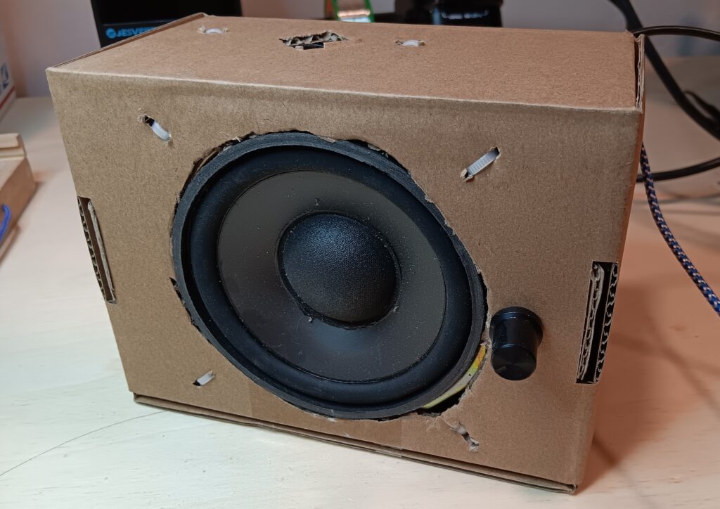 Front view of a large TV speaker built into the front of a cardboard box and a smaller speaker built into the top. The speakers are held in place with zip ties. There is a black volume control knob to the right of the front speaker. A blue-cloth wire and a black wire exit the top right rear corner of the box.